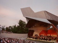 © Lisa Edi Blick auf den modernen Wolkenturm in Grafenegg bei Sonnenuntergang, mit einem Orchester auf der Bühne und zahlreichen Zuschauer:innen im Freiluftbereich.