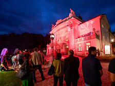 © Helge Bauer Menschen versammeln sich am Abend vor einem hellrot beleuchteten Theatergebäude. Das Gebäude ist kunstvoll verziert, und die Fassade wird durch farbiges Licht eindrucksvoll in Szene gesetzt. Der Himmel ist tiefblau, und die Besucher stehen auf dem gepflasterten Vorplatz und beobachten das Geschehen.