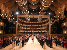 © Katharina Schiffl Festlich beleuchteter Opern- oder Ballsaal mit mehreren Kronleuchtern und voll besetzten Rängen. Auf dem Parkett tanzen zahlreiche Paare in Abendkleidern und Anzügen in einer symmetrischen Formation, umgeben von Publikum.