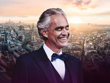 © Leutgeb Entertainment Ein Mann im Smoking mit Fliege lächelt vor einer Stadtansicht bei Sonnenuntergang.