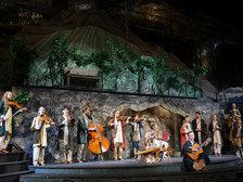 © Franz Neumayr Junge Musiker:innen in traditionellen Gewändern musizieren mit Geigen, Kontrabass, Zither, Akkordeon und Gitarre vor einer Bühnenkulisse mit Weinreben und Bergen beim Salzburger Adventsingen.