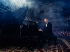 © Christian Barz Ein Pianist sitzt in einem eleganten Konzertsaal an einem schwarzen Flügel und spielt konzentriert. Um ihn herum liegt leichter Bühnennebel, der die Atmosphäre geheimnisvoll wirken lässt. Der Saal ist leer, das Licht fällt weich auf die roten Sitzreihen und die kunstvoll gestalteten Wände im Hintergrund.
