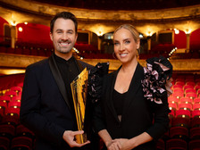 @ ORF / Hans Leitner Ein Mann und eine Frau in eleganter schwarzer Abendgarderobe stehen lächelnd in einem prachtvollen Theater mit rotem Samtinterieur. Der Mann hält eine goldene Trophäe in den Händen.